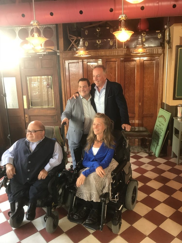 Dans un décor de restaurant, deux hommes debout derrière deux personnes en fauteuil roulant posent pour la photo.